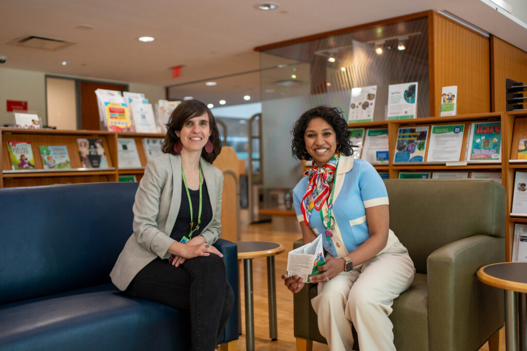 Two adults sit on arm chairs in the library, surrounded by shelves of books and pamphlets.