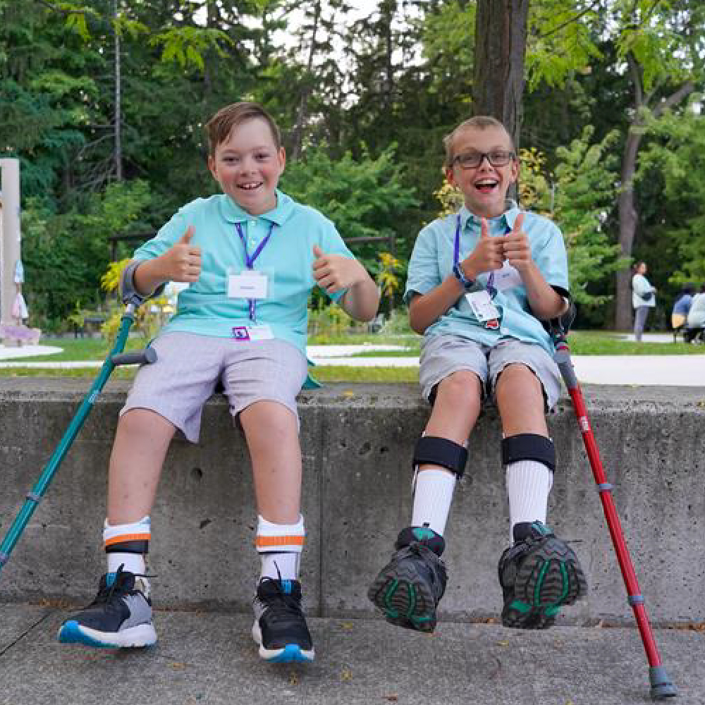 Two young boys sit outside in front of trees, smiling with their thumbs up