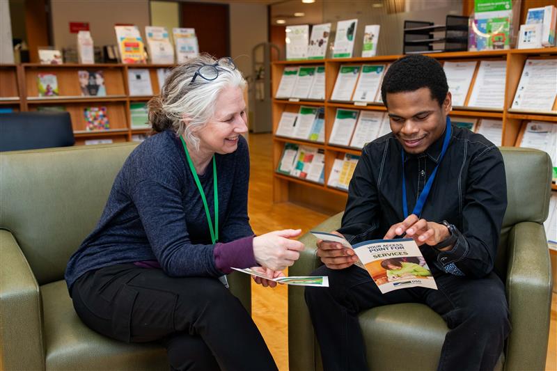 A woman and a young man sit together in a library and read a pamphlet