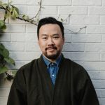 Headshot – Genuine Lam An adult wears a suit in front of a white brick wall