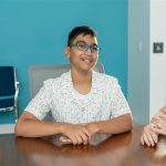Rehan-2 A young boy smiles while sitting in a board room in front of a blue wall