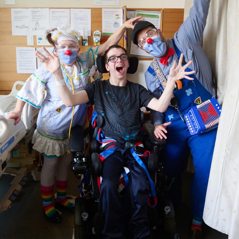 about-grid001 A young person using a wheelchair smiles and waves their hands with two therapeutic clowns