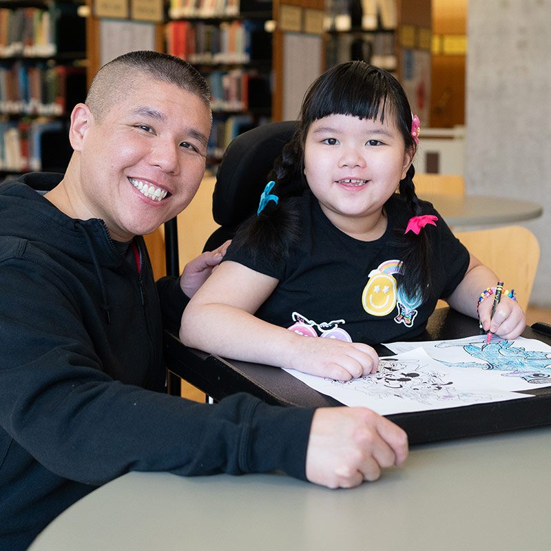 An adult and child colour a picture while sitting in a library