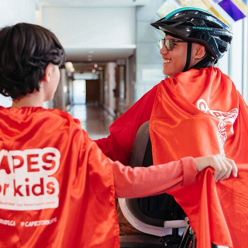 Two kids race each other in the hospital hallway wearing red capes